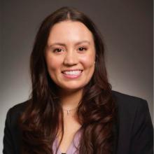 A person in a black blazer and purple blouse poses in front of a grey background.