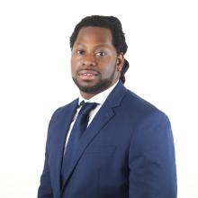 A person in a blue suit stands in front of a white background.