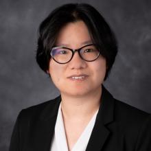 A person in a black blazer and glasses stands against a grey background.