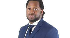 A man in a blue suit stands in front of a white background