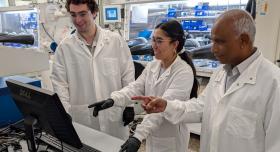 Three researchers wear white lab coats and stand in front of a computer monitor in a lab.