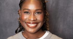 A person in a grey sweater poses for a headshot in front of a grey background.