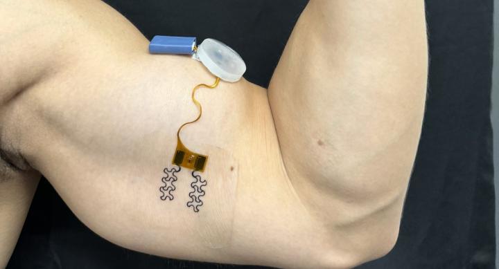 A man flexes his bicep, on which he wears a sensor made up of a small blue battery, a plastic cap, and an orange wire taped below the muscle.