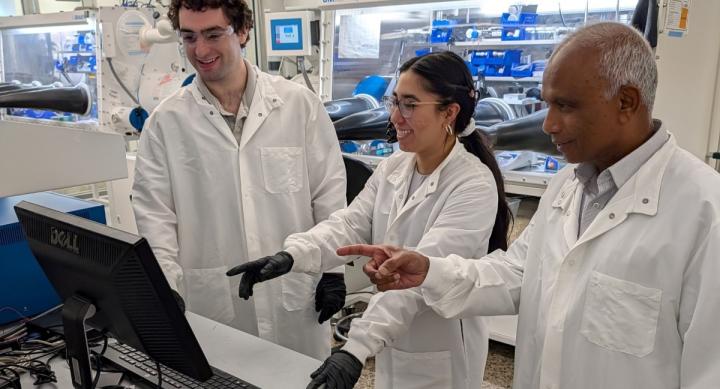 Three researchers wear white lab coats and stand in front of a computer monitor in a lab.