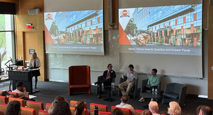 Three people sit in chairs under a powerpoint presentation and deliver a panel discussion to a lecture hall.